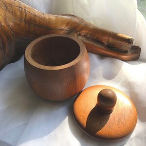 以下が含まれることがあります： 木製の丸い容器と蓋。どちらも濃い茶色です。蓋には小さなつまみが付いています。容器と蓋は木製で、自然な木目が見えます。容器の直径は約10cmです。
