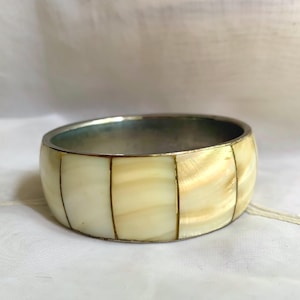 May include: A chunky bracelet in ivory and gold tones, with a silver-coloured interior. The bracelet is divided into rectangular segments. The jewellery item is likely made from a natural material, possibly shell or stone.