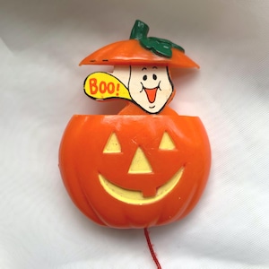 May include: Orange plastic Halloween decoration featuring a smiling jack-o'-lantern with triangular eyes and a smiling mouth. A ghost figure pops out of the pumpkin, holding a speech bubble that says "Boo!". The pumpkin has a green stem and a red string.