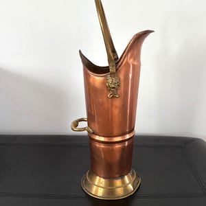 May include: A copper and brass coal scuttle with a handle. The scuttle has a tall, cylindrical body with a flared top and a brass base. A brass lion's head adorns the front. The handle is made of brass.