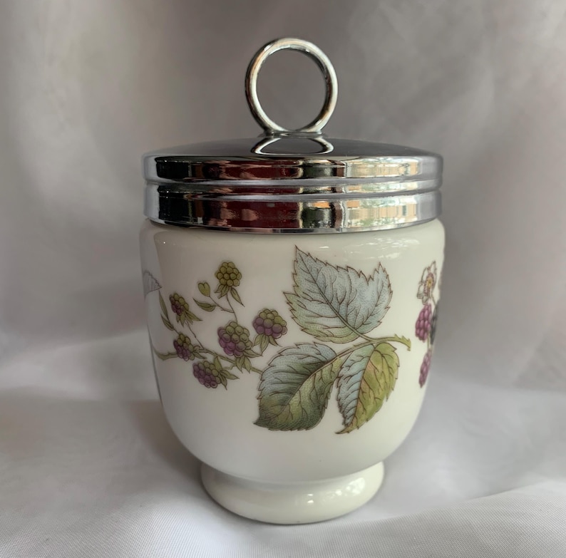 K&ouml;nnte beinhalten: Ein wei&szlig;er Eierbecher aus Keramik mit einem silbernen Deckel und einem Blumendesign mit Brombeeren und Bl&auml;ttern.