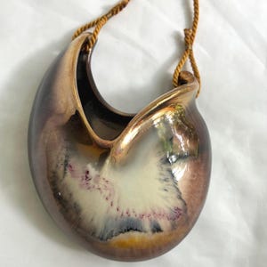 May include: A crescent-shaped ceramic wall pocket planter with a glossy metallic bronze and brown glaze. The center features a cream-colored splash pattern with subtle purple and black accents. It hangs from a twisted golden-brown cord.