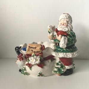 May include: A ceramic Santa Claus figurine with a green and white coat, holding a red bird and a white bird. The figurine is part of a container with the words "Tweet Treats" on a wooden sign. The container is decorated with a red ribbon and white flowers.