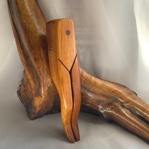May include: A hand-carved wooden nutcracker featuring a smooth, light brown finish with a distinct grain pattern. The tool has a vertical, elongated shape with a small circular inlay near the top and a central split design for cracking nuts.