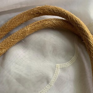 Puede incluir: Primer plano de dos asas tejidas de color beige, probablemente de un bolso o cartera. Las asas son gruesas y texturizadas, con un patr&oacute;n tejido apretado. El fondo es una tela blanca suave.