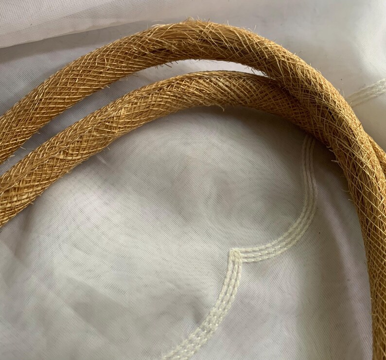 Puede incluir: Primer plano de dos asas tejidas de color beige, probablemente de un bolso o cartera. Las asas son gruesas y texturizadas, con un patr&oacute;n tejido apretado. El fondo es una tela blanca suave.