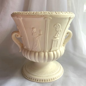 May include: Off-white decorative urn with relief carvings of classical figures and columns. The urn has two curved handles and a pedestal base. The rim features a decorative border. The design is reminiscent of ancient Greek or Roman art.