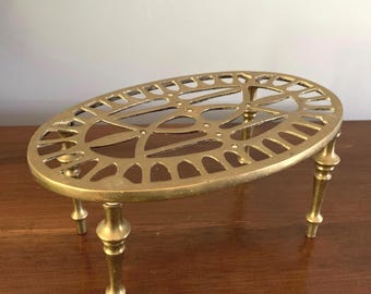 May include: A brass trivet with an oval, openwork design. The trivet has four ornate legs and a decorative pattern on the top surface. The color is a warm gold, and the trivet appears to be made of metal.