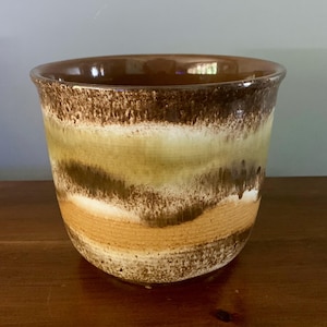 May include: A ceramic plant pot with a wide, slightly flared rim. The exterior features horizontal bands of colour, including brown, tan, white, and olive green. The pot has a textured, organic appearance, suggesting a handmade or vintage aesthetic.