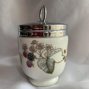 K&ouml;nnte beinhalten: Ein wei&szlig;er Keramik-Eierbecher mit einem silbernen Deckel und einem Blumendesign mit Brombeeren und wei&szlig;en Bl&uuml;ten.
