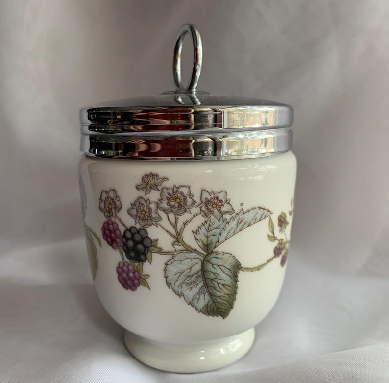 K&ouml;nnte beinhalten: Ein wei&szlig;er Keramik-Eierbecher mit einem silbernen Deckel und einem Blumendesign mit Brombeeren und wei&szlig;en Bl&uuml;ten.