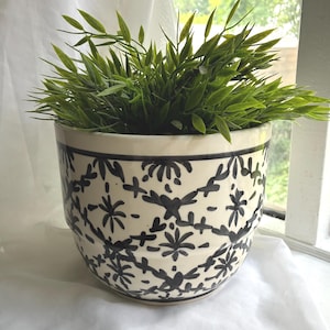 May include: A ceramic planter with a cream-coloured base and a black hand-painted floral pattern featuring repeating diamond shapes and star-like motifs. The pot contains a lush, green artificial plant with pointed leaves filling the top.