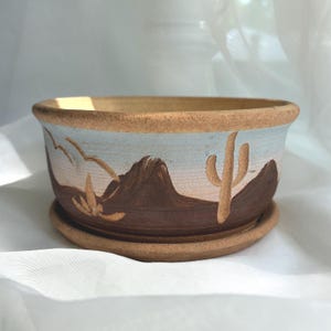 May include: A round ceramic planter with a matching saucer, featuring a hand-painted desert landscape. The design includes brown mountains and a saguaro cactus against a pale blue and white sky, with a natural, unglazed tan clay rim and base.