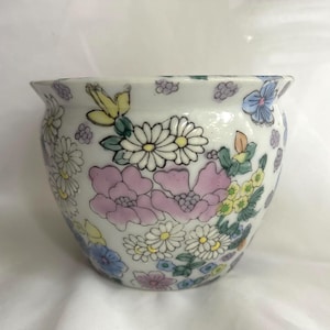 May include: A white ceramic planter with a floral design. The planter features pink, yellow, and blue flowers, along with green leaves. The flowers are painted on the exterior of the planter.