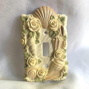 May include: Off-white light switch cover with seashell and rose embellishments. The rectangular cover features a central switch opening, surrounded by sculpted seashells, roses, and green leaves. A large seashell tops the design.
