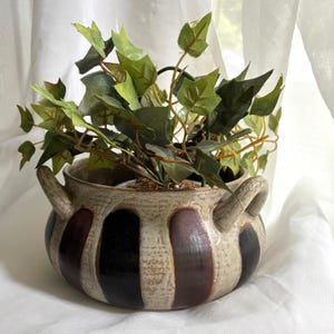 May include: A ceramic planter with two side handles featuring a speckled beige base and vertical stripes in alternating shades of dark brown and black. The pot contains a faux green ivy plant with pointed leaves extending from the top opening.