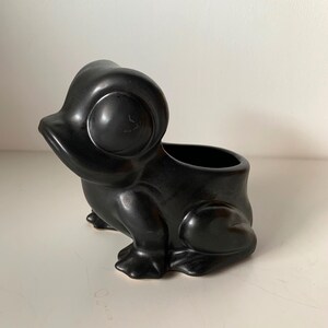 May include: A matte black ceramic planter shaped like a stylised frog. The frog figure features large, rounded eyes, a wide mouth, and a hollowed-out back section designed to hold small items or plants. The surface has a smooth, uniform finish.