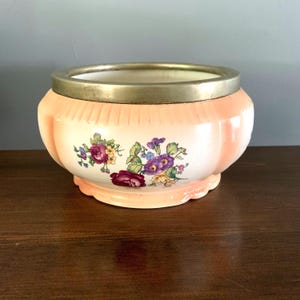 May include: A peach-colored ceramic planter with a scalloped base and a gold-toned rim. The planter features a floral design with purple, pink, and yellow flowers. The planter is a decorative item.