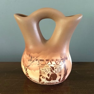 Peut inclure: Un vase en céramique marron avec une anse unique et un motif décoratif. Le vase présente une ouverture centrale et deux becs. La partie inférieure est ornée d'une scène gravée détaillée dans des tons bruns et crème.