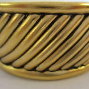 May include: A gold-colored ring with a braided design. The ring is made of metal and has a black accent.