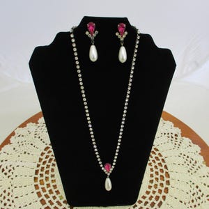 May include: A long, silver rhinestone necklace with a pink teardrop pendant and a pearl drop. Matching earrings with pink teardrop stones and pearl drops.