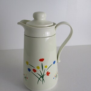 May include: White thermal carafe with a floral design in red, yellow, and blue. The carafe has a lid with a handle and a separate handle on the side.