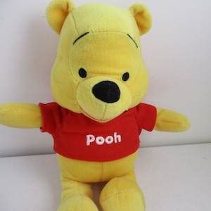 May include: A yellow plush Winnie the Pooh bear wearing a red shirt with the word "Pooh" embroidered in white.