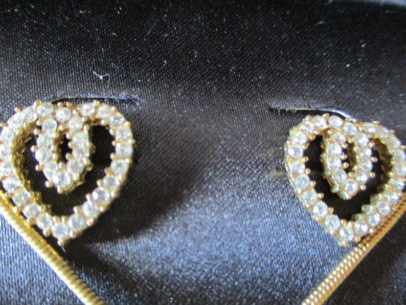 Puede incluir: Un par de pendientes de tono dorado con un dise&ntilde;o de doble coraz&oacute;n, cada coraz&oacute;n est&aacute; incrustado con diamantes de imitaci&oacute;n transparentes.