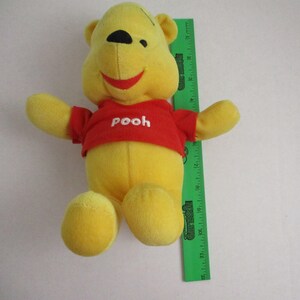 May include: A yellow plush Winnie the Pooh bear wearing a red shirt with the word "pooh" printed on it. The bear is sitting on a white surface.