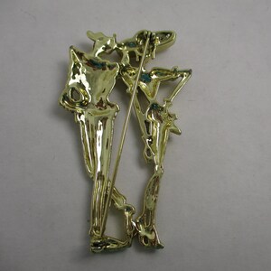 May include: Gold-toned metal brooch featuring a skeletal figure holding a staff. The brooch is detailed with intricate lines and curves.