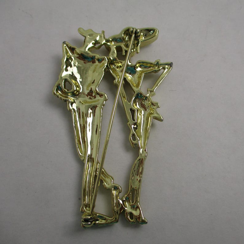 May include: Gold-toned metal brooch featuring a skeletal figure holding a staff. The brooch is detailed with intricate lines and curves.