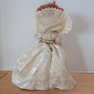 Puede incluir: Una mu&ntilde;eca de porcelana que lleva un vestido blanco con ribete de encaje y un cintur&oacute;n dorado. La mu&ntilde;eca tiene el pelo rojo y lleva una cofia de encaje blanco.