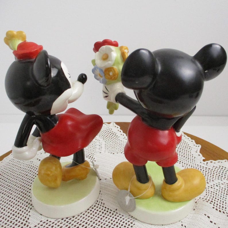May include: Two ceramic figurines of Mickey and Minnie Mouse. Mickey, in red shorts, presents a bouquet to Minnie, who wears a red skirt and hat. Both figures have black bodies, white gloves, and stand on a light green base.