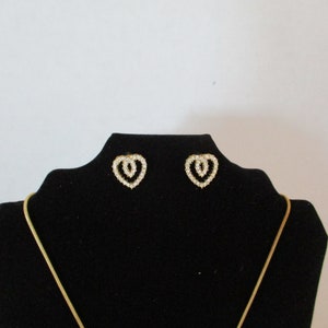Puede incluir: Un par de pendientes de coraz&oacute;n de oro con diamantes de imitaci&oacute;n transparentes en un expositor de terciopelo negro. Tambi&eacute;n hay un collar de cadena de oro en el expositor.