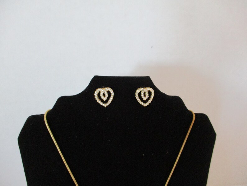 Puede incluir: Un par de pendientes de coraz&oacute;n de oro con diamantes de imitaci&oacute;n transparentes en un expositor de terciopelo negro. Tambi&eacute;n hay un collar de cadena de oro en el expositor.