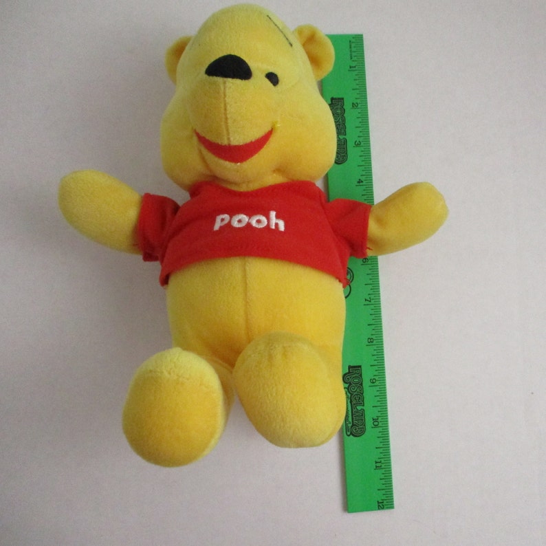 May include: A yellow plush Winnie the Pooh bear wearing a red shirt with the word "Pooh" embroidered on it. The bear is sitting with its legs outstretched.