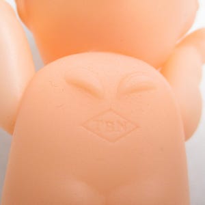 May include: A close-up of a vintage, peach-colored baby doll. The doll's back is visible, with a molded design of a diaper area and a diamond-shaped mark with the letters "TBN". The doll's arms and head are also visible.