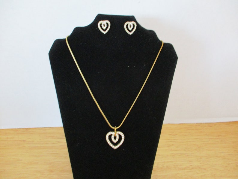 Puede incluir: Un collar de cadena dorada con un colgante de coraz&oacute;n doble y pendientes a juego. Los corazones est&aacute;n engastados con diamantes de imitaci&oacute;n transparentes.