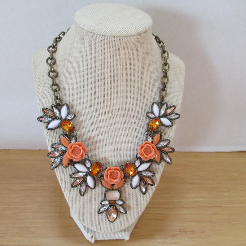 May include: A statement necklace with a floral design featuring orange and white flowers, clear and amber rhinestones, and a gold chain.