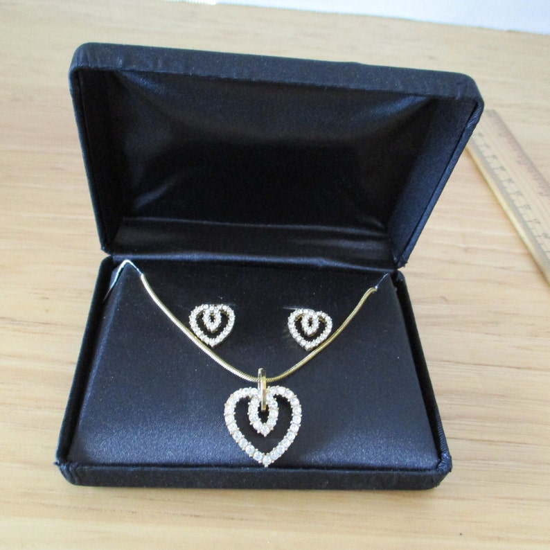 Puede incluir: Un conjunto de collar y pendientes de coraz&oacute;n de oro con detalles de cristal transparente, presentado en una caja de terciopelo negro.