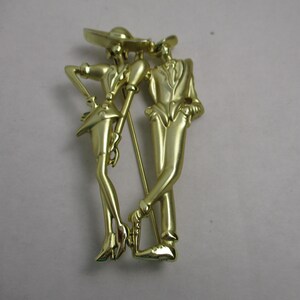 May include: Gold-toned metal brooch depicting a stylized couple dancing. The woman is wearing a hat and a short skirt, while the man is wearing a suit. The brooch is in a vintage style.
