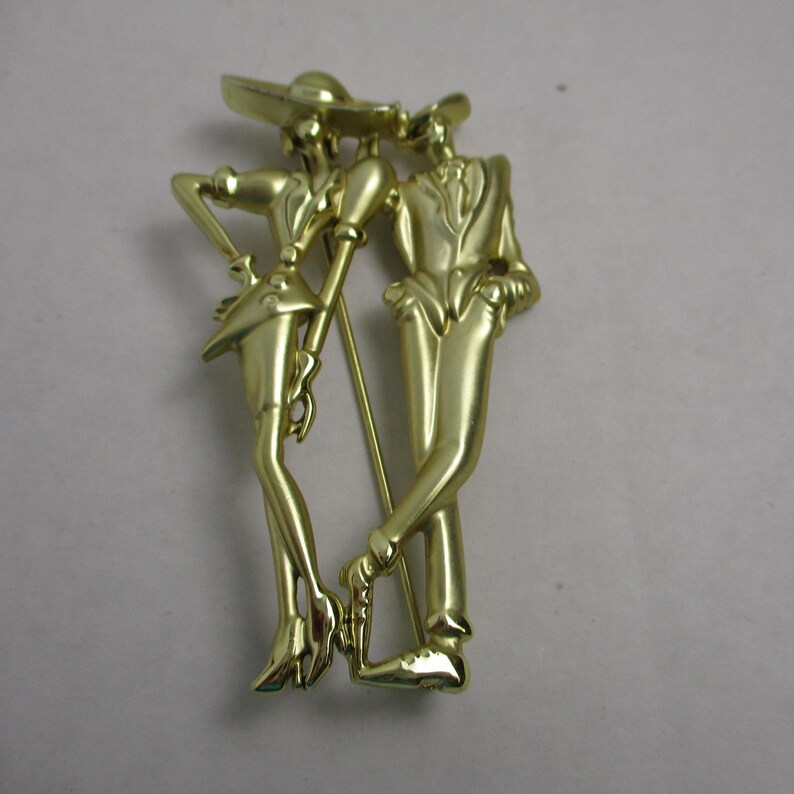 May include: Gold-toned metal brooch depicting a stylized couple dancing. The woman is wearing a hat and a short skirt, while the man is wearing a suit. The brooch is in a vintage style.