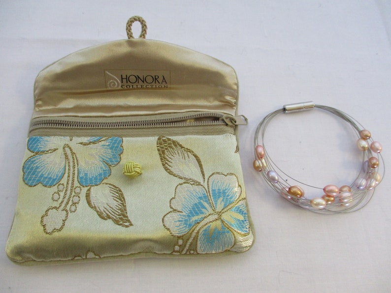 Op de afbeelding: Een gouden satijnen tasje met een bloemenpatroon en de tekst "Honora Collection" erin. Het tasje is open en een zilveren draad armband met roze en paarse parels ligt ernaast.