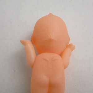 May include: A peach-colored plastic doll, viewed from the back, with arms outstretched. The doll has a rounded head and body, with a small mark on the back. The doll's arms are slightly bent, and the hands are open.