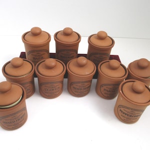 May include: A collection of ten cylindrical terracotta spice jars with matching lids. Each jar features a dark brown printed label with the text The Original Suffolk Collection and specific spice names including Basil, Rosemary, Chives, Garlic, Cloves, Mixed Herbs, Parsley, and Peppercorns.