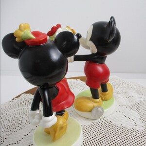 May include: Figurines of Mickey and Minnie Mouse in a romantic pose. Mickey is wearing red shorts, yellow shoes, and black ears. Minnie is wearing a red dress, yellow shoes, and a red hat with a yellow flower. Both are on a green base.
