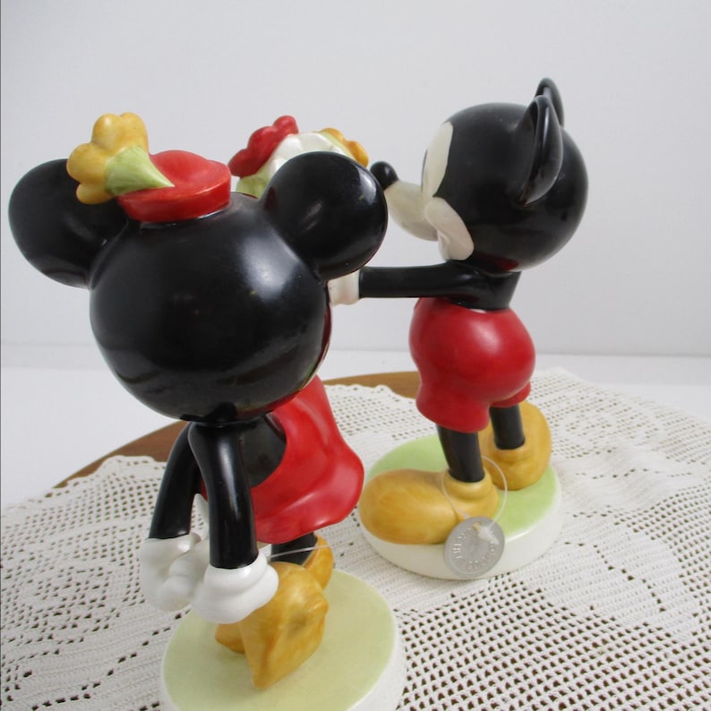 May include: Figurines of Mickey and Minnie Mouse in a romantic pose. Mickey is wearing red shorts, yellow shoes, and black ears. Minnie is wearing a red dress, yellow shoes, and a red hat with a yellow flower. Both are on a green base.