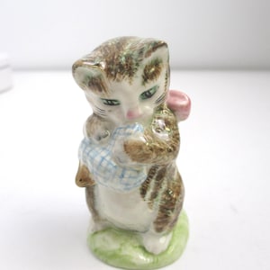 May include: A ceramic figurine of a brown tabby kitten standing on a green base. The kitten has green eyes, a pink nose, and is holding a blue and white checkered bundle in its front paws. A small pink object is visible behind the kitten's head.
