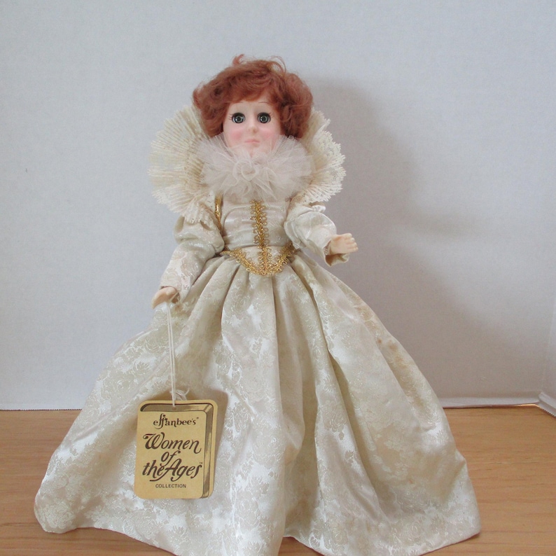 Puede incluir: Una mu&ntilde;eca de porcelana con un vestido blanco con ribetes dorados y un cuello de encaje blanco. La mu&ntilde;eca tiene el pelo rojo y los ojos marrones. La mu&ntilde;eca sostiene una etiqueta que dice "Effanbee's Women of the Ages Collection".