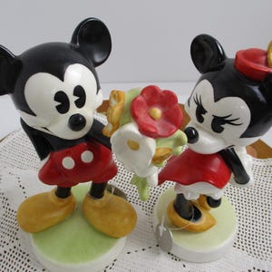 May include: Two ceramic figurines of Mickey and Minnie Mouse. Mickey, in red shorts and yellow shoes, holds a bouquet. Minnie, in a red dress and hat, stands nearby. Both figures have black ears and white faces, set on green bases.